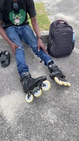 A man sits on a concrete curb wearing black roller blades, blue jeans, a black t-shirt with a graphic, a black wristwatch, and a black beanie.  His feet are spread apart and forward. He is talking to someone. Next to his left foot are a pair of black sandals. Behind the man and to his right is a black backpack and a bottle of water. The concrete he sits on is gray. Behind the curb is grass.