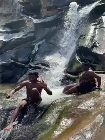 Two people are at the bottom of a waterfall. One person is seated on a rock while the other is crouched down and reaching toward the water. The water is clear and splashing around them. The surrounding rocks are dark and mossy. The video is shot from a low angle.