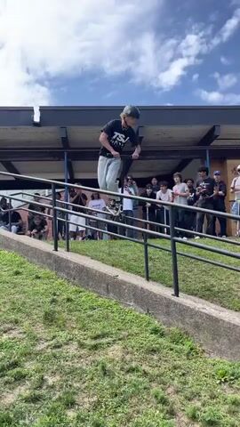 The video shows a young person on a scooter performing a trick on a ramp. They are wearing a helmet and a black t-shirt with white writing on it. They are also wearing light-colored pants. The ramp is made of concrete and metal. There is a crowd of people watching the scooter rider, some of whom are holding phones to record the event. The scene appears to be outdoors on a sunny day, with trees and a building in the background. The crowd is standing behind a railing. The scooter rider is in mid-air, performing a jump. The crowd is looking up at the rider.
