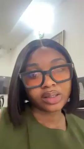 A woman with dark hair cut in a short bob is shown in a close-up, looking directly at the camera. She is wearing large, dark-framed glasses with orange-tinted lenses. She is also wearing an olive green top. The background includes a white ceiling, a framed picture on the wall, and a glimpse of a television and an air conditioning unit. She has a neutral expression and slight movements of her mouth. The camera angle is slightly from below.