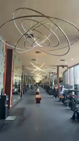 The video shows the interior of a fitness center or gym, viewed from one end looking down a long corridor. Along both sides of the corridor are mirrored walls and individual exercise stations with rowing machines and other equipment. Several people are present, standing or working out at the stations. The most striking feature of the space is a series of large, elaborate chandeliers hanging from the ceiling, evenly spaced down the length of the corridor. These chandeliers are made up of multiple intersecting gold-colored rings with integrated lighting, creating a complex and modern design. The floor is a dark color, and wooden benches are placed along the corridor for seating. Natural light enters from a window on the right side of the frame.