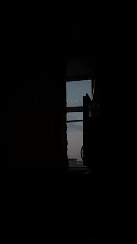 A dark room with curtains drawn on the left and right. In the center, a window is visible. The window pane appears to be cracked in a few places. Through the window, a dusky sky with clouds is visible. The silhouette of what appears to be a fan is visible on a surface below the window.