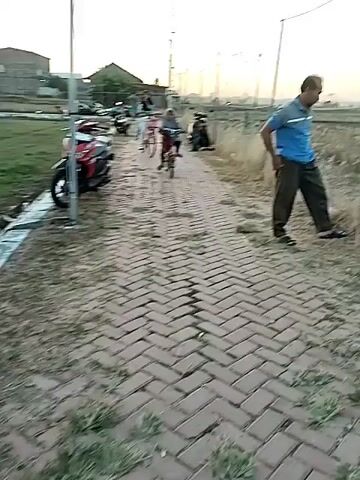 The video shows a group of people riding bicycles and motorbikes along a brick-paved path. The sun is low in the sky, casting a warm glow over the scene. In the background, there are buildings, trees, and a net fence. The people appear to be enjoying a leisurely ride, and the overall atmosphere is peaceful and relaxed.