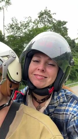A video captures two people on a motorcycle, both wearing helmets. The person in front, whose face is partially visible, wears a white and cream helmet with green and white accents. The person behind, who is the primary focus, wears a black helmet with pink accents and a blue and white plaid shirt. They are smiling and looking at the camera. The background shows a road lined with trees and greenery, indicating an outdoor, possibly rural, setting. Another motorcycle with two riders is visible further down the road. The video appears to be a selfie video taken while riding.
