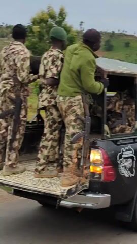 The video shows the rear of a black pickup truck traveling on an asphalt road. The truck has a metal cage structure built into the truck bed, where several people in camouflage uniforms are seated. Three additional people, also in camouflage uniforms and holding rifles, stand on the truck's open tailgate. The truck's rear side is labeled "OPERATION FUSHIN-KADA KADUNA STATE GOVERNMENT." The environment includes a green, hilly landscape in the background. The sky appears overcast.