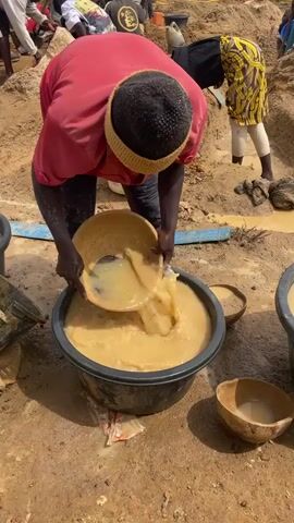 The video shows a person in a pink shirt and black pants, wearing a black cap with a tan brim, leaning over a large, grey tub. The person is pouring a tan-colored liquid from a round, wooden bowl into the tub. The tub is filled with a similar tan-colored liquid, and there are other buckets and wooden bowls around the tub on the ground. The ground appears to be sandy and muddy. Other people are visible in the background, some working and some standing around. One person in the background is wearing a yellow and black patterned top with white pants.