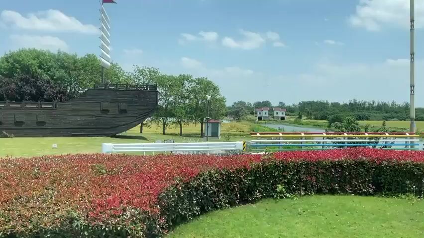 The camera moves across a rural, sunny, outdoor scene. A ship-shaped sculpture is visible on the left. A small stream or narrow lake is in the center, with a small white house with a red roof visible in the distance. A multi-colored pipe and railing structure runs along the water's edge.  Green and red ground cover vegetation is in the foreground.  Power lines and sparse trees are also visible.
