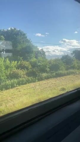 The video shows a view from inside a moving vehicle looking out a window. The scene outside is a rural landscape with green trees and fields under a bright blue sky with scattered white clouds. The ground is covered in grass, some of which appears to have been recently mowed. There are utility poles and what appear to be electrical boxes along the side of the road. In the distance, there are some buildings and a car. The camera pans slightly to the right, showing more of the scenery as the vehicle continues to move.