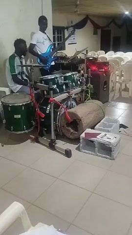 The video captures a scene inside a room with several rows of white chairs arranged as if for an event or gathering. The room is brightly lit, and the floor is covered with light-colored tiles. Several musicians are present, setting up equipment or preparing to perform. 

Initially, the focus is on a man playing an electric guitar, then pans to a man sitting at an electronic keyboard. A woman with a towel draped over her head is also visible in the shot, seated in the foreground, and the camera subsequently shifts to a man smiling and holding several microphones.

Scattered around the musicians are various musical instruments, including drums, congas, and a speaker. There are also some rolled-up materials and other equipment on the floor.