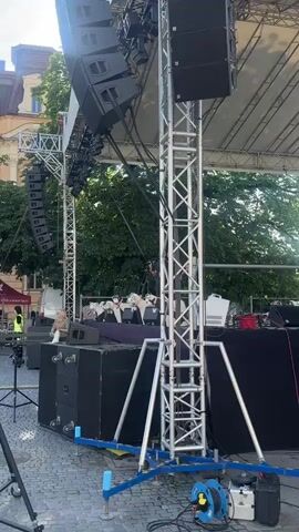 A video begins with a close-up shot of a stage setup. Large speakers are visible, as is a metal truss structure holding up a sound system. The camera pans slightly to reveal a row of stuffed animals, possibly puppets, positioned on the stage. Each animal appears to be a dog, with distinct markings and collars. Behind the animals, several microphones on stands are set up. Two individuals, presumed to be performers or crew members, are visible on the stage. One person is holding a microphone and speaking, while another stands nearby. The background consists of trees and a building, suggesting an outdoor event. The lighting indicates it is daytime, possibly late afternoon. The camera continues to capture the scene, focusing on the stage and its elements.