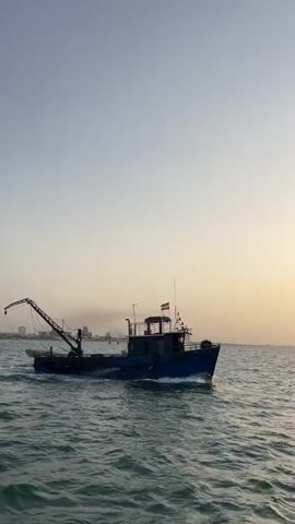 A blue fishing boat is moving through the water under a clear sky. The boat has a crane on its deck and emits smoke from its funnel. In the background, a city skyline is visible on the horizon. The sun appears to be setting, casting a warm glow on the scene.