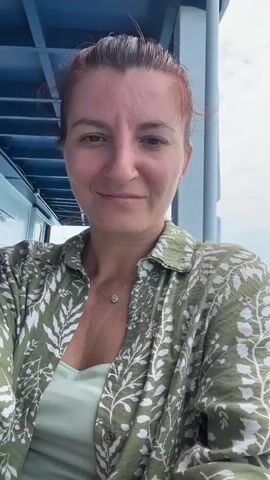 A woman with reddish-brown hair tied back, wearing a green leafy-patterned collared shirt over a light green top and a delicate necklace, records a selfie video on a boat. She has a nose piercing and is smiling gently at the camera. The background shows open water with a slight chop, indicating movement. A light blue structure of the boat is visible to her left. In the distance, a faint coastline or island can be seen under an overcast sky. The setting suggests a daytime boat journey in a location like Ao Wiang Wan, Thailand. The woman appears calm and content.