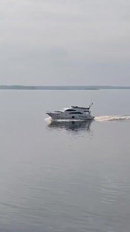 The video shows a white yacht moving across a calm body of water. The sky is overcast and the water is a grey color. Land is visible in the far distance. The yacht leaves a trail of white water behind it.