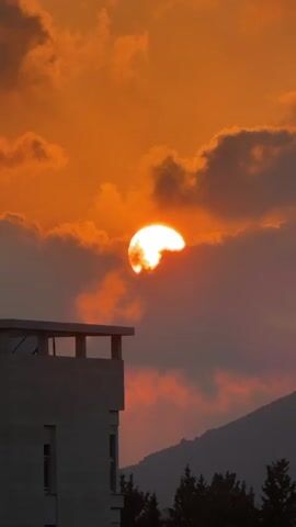The video captures a sunset scene. The sun, partially obscured by clouds, emits a bright orange glow. In the foreground, a light-colored building with a balcony is visible. Behind the building, silhouetted trees and mountains form the background. The sky is a vibrant orange with scattered clouds, and the overall atmosphere suggests the end of the day.