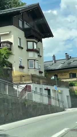 The video shows a view from a moving vehicle on a sunny day with a partly cloudy sky. The scene is a street in a town with buildings and trees. The initial part of the video shows parts of buildings and a blue van driving on the road. As the video progresses, more buildings are visible, including one with "APARTMENT Harrer" written on it. Several other vehicles, including cars and vans, are seen driving on the road. In the later part of the video, a white building and a set of train tracks are visible. Mountains are present in the background.