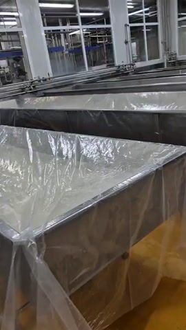 The video shows a factory floor with multiple large stainless steel vats, some covered with clear plastic sheeting. A man in a white shirt and vest is visible in the background, operating a yellow hose, likely cleaning or rinsing equipment. The floor is a light yellow color and appears wet in some areas, indicating recent cleaning or an ongoing process. The overall environment suggests a food processing or manufacturing facility.