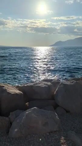 The video shows a still shot of a body of water, likely the sea, with a distant mountainous coastline. The sun is high in the sky, casting a shimmering reflection on the water. In the foreground, there are several large rocks, with gravelly ground beneath them. The sky has some scattered clouds. The camera appears to be stationary.