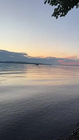 The video shows a tranquil scene of a body of water, likely a lake or sea, at sunset. The sky displays a gradient of colors, from pale blue at the top to soft oranges and pinks near the horizon, where clouds are visible. Ripples on the water's surface catch the fading light, creating a shimmering effect. In the distance, a small boat is visible on the water. The foreground shows a shoreline with pebbles. The overall atmosphere is peaceful and serene.