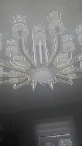 The video shows a close-up, low-angle shot of a large, ornate chandelier hanging from a white ceiling. The chandelier is made of crystal and gold-colored metal, with multiple tiers and arms holding individual light fixtures. The overall scene is dimly lit, with the chandelier itself being the brightest element. The focus is on the intricate details of the chandelier's design, including its faceted crystal components and metallic accents. The background is largely out of focus, but hints of a room with a window and potentially an air conditioning unit are visible at the bottom of the frame. There is no visible text within the video. The time of day and weather are not discernible. The atmosphere is still and quiet, focusing solely on the opulent decoration.