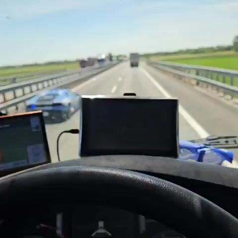 A video shows the view from the cab of a large truck on a highway. The road is relatively straight, with several tractor-trailers ahead. A blue Lamborghini sports car, marked as POLIZIA, is in the lane to the left, moving with its blue lights activated. The landscape includes a median barrier and green fields adjacent to the highway. The day appears bright with a clear sky. The camera perspective is stable, focusing on the vehicles ahead.