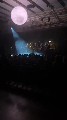 The scene is an indoor concert hall where an orchestra is performing for a seated audience. The stage is dimly lit with a spotlight focused on a woman in a long dress playing the violin. Smoke or fog effects are used on stage, partially obscuring the performers. Above the stage, there is a large, illuminated sphere resembling the moon. The audience is visible as dark silhouettes in the foreground. The ceiling structure with lights and scaffolding is visible above the stage.
