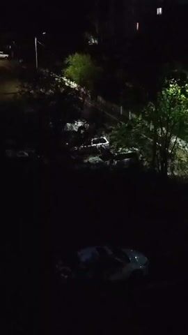The video shows a nighttime scene from an elevated viewpoint. A dimly lit area contains several parked cars. One white car is visible in the foreground, and other vehicles are further back, partially obscured by darkness and foliage. Trees and bushes are scattered throughout the scene. In the background, a building with illuminated windows suggests a residential area. A street light is also visible, casting a faint glow on the surroundings. The overall impression is a quiet, dimly lit urban or suburban landscape at night.