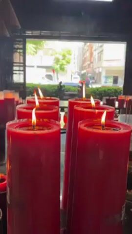 The video shows several thick, red, cylindrical candles burning with small flames on a reflective surface.  There are more candles and other objects in the background. Behind the candles, a window with bars reveals an outdoor scene including a building, foliage, and vehicles. The lighting is mixed, with the flames providing a warm glow against the daylight outside.