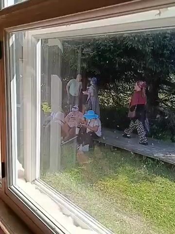 The video begins by looking through a window at an outdoor scene. Inside the frame, there are at least four people visible. One person wearing a blue hat and a light-colored shirt is sitting on the ground and appears to be engaged in an activity. To the left, another person is standing and partially obscured. Further back, two other individuals are visible, one wearing a headscarf. They are situated on a wooden walkway that leads away from the viewer. Behind them, dense green trees are present. The camera then pans slightly, revealing more of the surroundings. A yellow sign on an easel is visible to the right of the walkway. The scene transitions to a wider view, still through the window, showing a grassy area in the foreground. Two individuals, an older man and woman, are seated on a bench, facing the camera and seemingly interacting. In the background, beyond a wooden fence, a group of people are gathered in a field with trees and buildings in the distance. The sky is clear and blue.