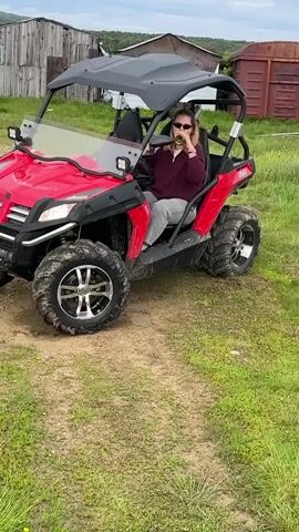 A woman sits in the driver's seat of a red all-terrain vehicle. She is wearing a maroon sweater, gray pants, and sunglasses. She holds a bottle with both hands, appearing to be opening it. The ATV is parked on a dirt path surrounded by grass. In the background, there are rustic wooden buildings and a rusted corrugated metal structure. The sky is cloudy.