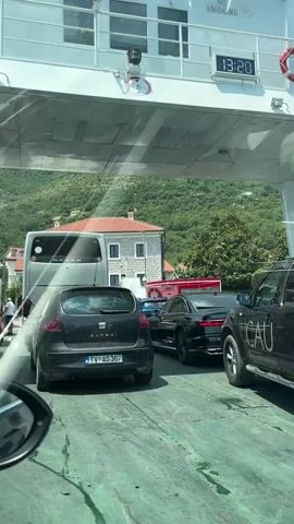 The video shows a queue of vehicles on a ferry. In the foreground, a black hatchback with the license plate TV AS 367 is visible from the rear. To its right, a black SUV with "CAU" written on its side is parked. Further ahead, a black sedan and a blue car are also lined up. A white bus is visible in the background, parked behind the black hatchback. Several buildings with red-tiled roofs are seen behind the vehicles, nestled against a lush green, mountainous backdrop. The sky appears bright, suggesting it's daytime. Above the vehicles, a white structure, presumably part of the ferry, has markings and text. One sign reads "NO SMOKING," and a digital clock displays "13:20." Another sign indicates a height limit of "4.5 m." A few people are visible in the scene, including one person standing near the blue car and another near the bus, possibly ferry personnel or passengers. The overall atmosphere appears calm, with vehicles patiently waiting. The setting is a ferry crossing, likely on a waterway given the mountainous terrain and buildings characteristic of coastal or lakeside towns in Montenegro, with the specific location being Đurići.