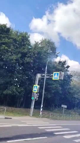 The video shows a view from a vehicle moving on a paved road. The sky is blue with white clouds. On the left side of the road, there are trees and a yellow sign. Further down the road, there are streetlights and a fence. In the distance, there is a white sign. A black car with a trailer is seen driving on the road. The video ends with a view of trees and fields on the right side of the road.