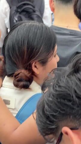The video captures various individuals from behind or in partial profile within a crowded public setting in Osaka, Japan. The camera frequently shifts focus between different people. One prominent subject is a woman with dark hair styled in a bun, wearing a light-colored jacket with blue straps, and a yellow earring. Her face is partially visible, showing a slight smile or an animated expression as if reacting to something. Other shots feature the backs of men's heads with diverse dark hairstyles, including short cuts and longer, wavy hair. Accessories such as glasses, simple necklaces, and backpacks are visible on some individuals. The scene portrays a bustling environment, suggesting people are moving or gathered in a busy street, station, or event area. No specific text is visible in the video.