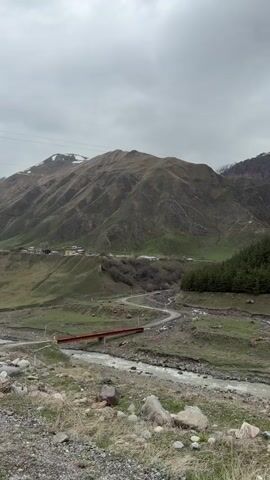 The camera faces a winding road leading to a small village at the foot of a large mountain on an overcast day. A rusty red bridge spans a rocky river that runs parallel to the road. Power lines stretch across the top of the frame. A small forest borders the road on the right, while sparse vegetation covers the rocky foreground. A white pole momentarily enters the frame.