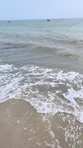 The video shows a view of a beach and the ocean. The sand is light brown and the water is a mix of green and grey. Small waves are rolling onto the shore, creating white foam. In the distance, there are a couple of small boats on the water. The sky is blue with some white clouds.