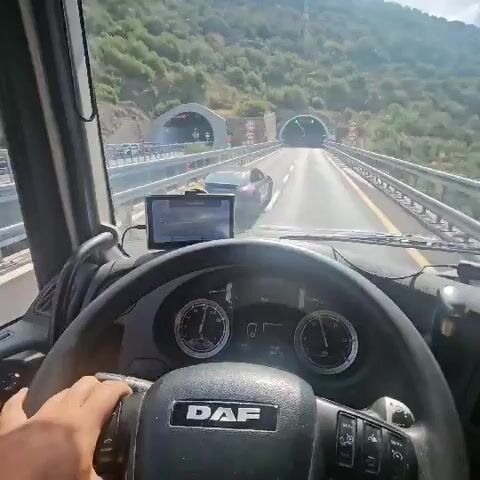 The video begins with a first-person perspective from the cab of a DAF truck. The camera is positioned behind the steering wheel, showing the driver's hand holding it. In front of the driver are the truck's dashboard instruments, including two large circular gauges and a digital display. A GPS device is mounted on the dashboard. The truck is on a highway, with a barrier to the right and a wooded hillside on the left. Ahead, there are two tunnels visible. As the truck approaches the first tunnel, a dark-colored car is seen ahead. The truck enters the first tunnel, and the lighting changes to the artificial illumination of the tunnel. The car continues to be visible in front. The video continues as the truck proceeds through the tunnel, with the camera maintaining the same perspective.