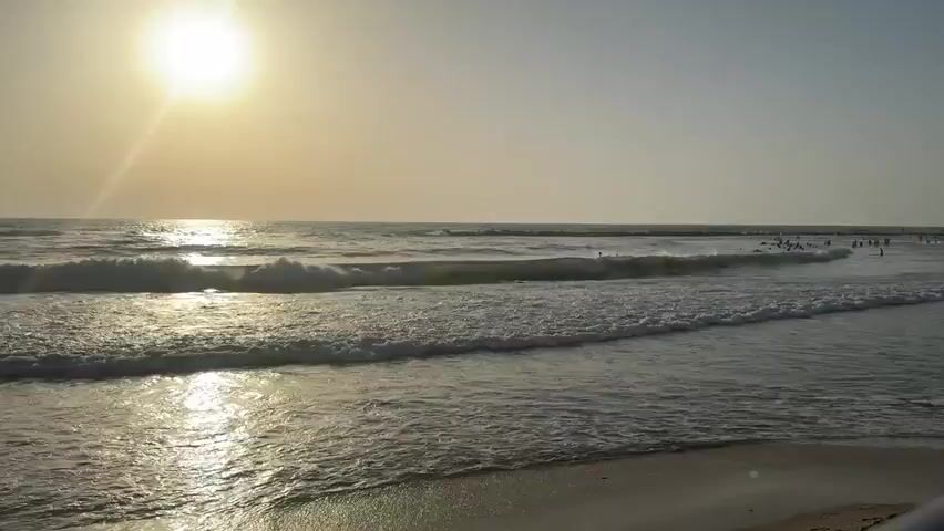 A wide shot shows a beach with the sun setting over the ocean. Gentle waves roll onto the sandy shore. In the distance, several people are visible in the water, some appearing to be swimming or wading. The sky is clear with a hazy orange glow near the sun. The overall mood is peaceful and serene.