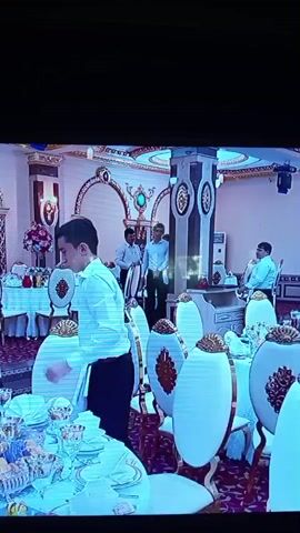 The video shows a large room with tables set for a formal event. The tables are covered with white tablecloths and decorated with elaborate floral centerpieces. There are numerous chairs with ornate white and gold designs. Several men, dressed in white shirts and dark pants, are moving around the room, seemingly preparing for the event. One man is adjusting items on a table, while others are talking or walking. The room itself is decorated with gold accents, arches, and decorative pillars, suggesting a luxurious setting. The lighting is bright, illuminating the scene.