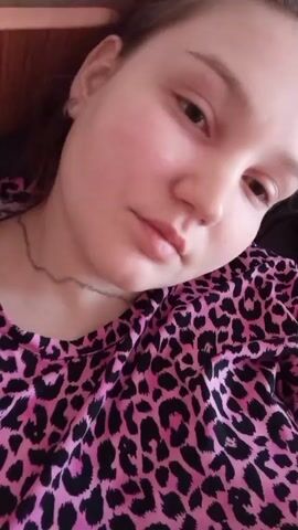 A young woman lies in bed next to a sleeping man. The woman is wearing a pink and black animal-print top. She blinks, looks toward the camera, and puckers her lips. The man in the background is shirtless and also appears to be asleep.