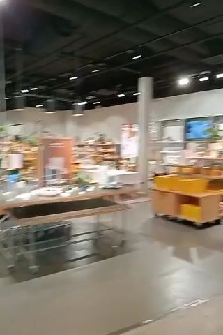 The video shows a wide shot of a shopping mall interior, with people walking in the distance. The camera moves forward, revealing a display of ceramic items in a store window on the left. The camera then pans to the right, showing more shops and shoppers. A prominent sign for "KULTAJOUSI" is visible on the right. The floor is tiled with a light wood-grain pattern. The lighting is bright, typical of a commercial indoor space.