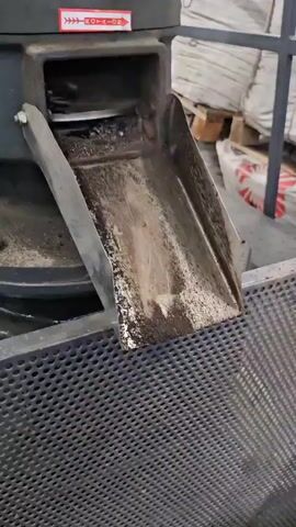 This video shows a close-up view of a conveyor belt or chute system where small, dark, cylindrical pellets are being dispensed. The pellets appear to be charcoal or a similar biomass material. They are falling from a metal chute onto a perforated metal screen, which forms a bin or container below. The pellets are accumulating in a large pile within the bin. The background is out of focus but appears to be an industrial setting with large white bags and pallets. The lighting is somewhat dim. There are no people visible in the video.