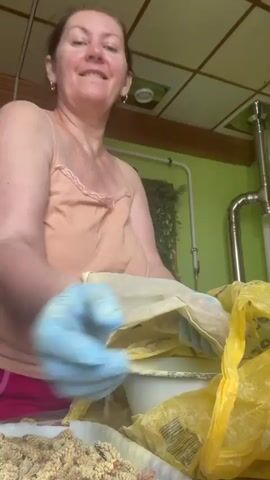 A middle-aged woman with light skin and brown hair, wearing a peach-colored tank top and bright pink shorts, is the main subject of the video. She is standing indoors in what appears to be a kitchen. She wears light blue gloves and is holding a small, off-white cloth bag filled with a granular substance. She is smiling and looking directly at the camera, seemingly talking. Behind her, a white bucket sits inside a large, yellow plastic bag, which contains several more of the filled cloth bags. To the left, a tray is visible, holding a large quantity of the same granular material that fills the bags. In the background, the kitchen walls are a light green color, and there is a drop ceiling. A stainless steel pipe runs along the wall.