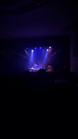 A dimly lit stage is illuminated by blue and yellow spotlights. Three figures are on stage, two of them are on either side of a table, and one is standing behind the table. The two figures on the sides are draped in white fabric, and the one in the center is wearing a white outfit. The figure in the center appears to be adjusting something on the table. The two figures on the sides are also adjusting the fabric. There is a drum set on the right side of the stage. The audience is visible in the foreground, silhouetted against the stage lights.