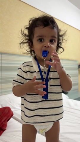 A young child, likely around one year old, stands on a bed wearing a striped polo shirt, a diaper, and a blue lanyard around their neck. They are holding a blue whistle in their hand and putting it in their mouth, then smiling at the camera, showing their teeth. The child has curly hair and dark eyes. A red cloth is visible in the lower left corner of the frame on the bed. The background consists of a patterned wall and a white bedsheet.