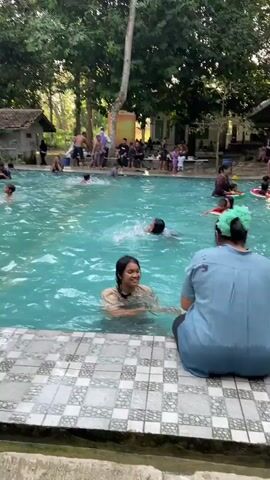 A group of children are swimming and playing in a clear, blue swimming pool surrounded by lush green trees. The pool has a tiled edge with a checkered pattern and a concrete walkway. In the background, there are small buildings that appear to be changing rooms or facilities. Several adults are also visible, some sitting by the pool and others in the water. The atmosphere appears to be relaxed and enjoyable, with the children actively engaged in their activities.