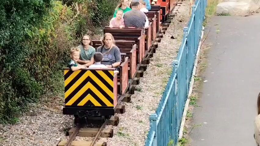 This is a video of a miniature train ride. The train is red and appears to be pulled by a locomotive. Several people, including adults and children, are riding in the open-air train cars. The train is on a track that runs alongside a path and a fence. There is lush green foliage on one side of the track. The train is moving forward and appears to be on an incline. The video is shaky at times, suggesting it was filmed by someone walking or moving with the train.