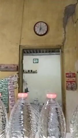 The video shows a row of clear plastic water bottles with pink caps lined up on a shelf. Behind the bottles, a doorway leads to another room where a clock is visible on the wall. Various items, possibly packages or wrappers, are hanging on the wall behind the bottles, and another shelf with items is visible further back. The overall environment appears to be a store or a storage area.