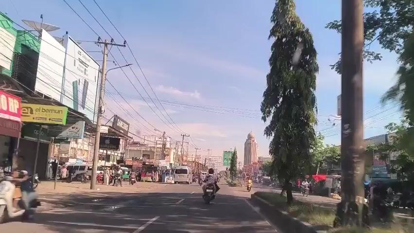 The video shows a street scene in what appears to be a city. Buildings line the left side of the street, with businesses displaying signs, including one for Samsung and another for Apple. Several people are walking along the sidewalk, and a few are standing near vehicles parked along the street. Vehicles, including cars, vans, and motorcycles, are driving on the road.  A tall tower is visible in the distance, possibly a minaret. Overhead power lines are strung across the sky. Trees are visible on the right side of the road, providing some greenery. The sky is blue with a few scattered clouds. The video is shot from the perspective of someone in a vehicle moving forward.