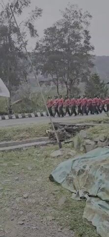 A group of people in matching red shirts and black pants are marching in formation on a paved road. They appear to be a marching band or a cadet group. There is an Indonesian flag on a pole to the left of the frame, waving in the wind. The background shows trees and hills, suggesting an outdoor setting, possibly in a rural or mountainous area. The ground in the foreground is covered with grass and some debris. The overall lighting suggests it is daytime, possibly overcast.