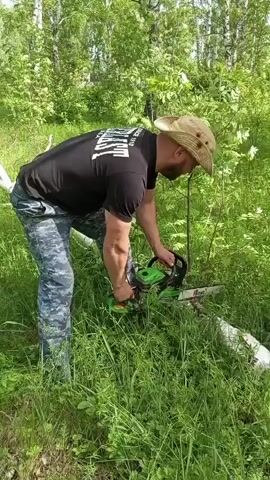 A man in a black shirt, camouflage pants, and a straw hat is cutting a fallen birch tree with a green and black chainsaw. The man is kneeling slightly with one knee on the ground and is positioned on the left of the frame, sawing the tree in the center. The scene takes place outdoors in a grassy, wooded area, with other trees and foliage visible in the background. The chainsaw produces sawdust as it cuts through the wood.