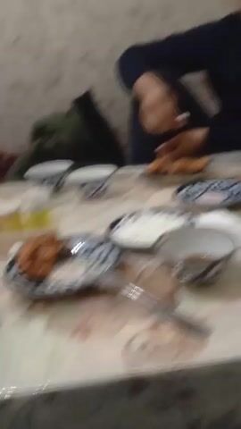 The video shows two young men at a table laden with food. The table is covered with a patterned tablecloth.  There are plates of food, small bowls, a bottle of Coca-Cola, and a teapot. The men are wearing dark clothing and appear to be eating. One man is picking at food with his hands, while the other man is sitting at the other end of the table. The background includes a wall with a light-colored patterned wallpaper.  The camera moves around the table, showing different angles of the scene.